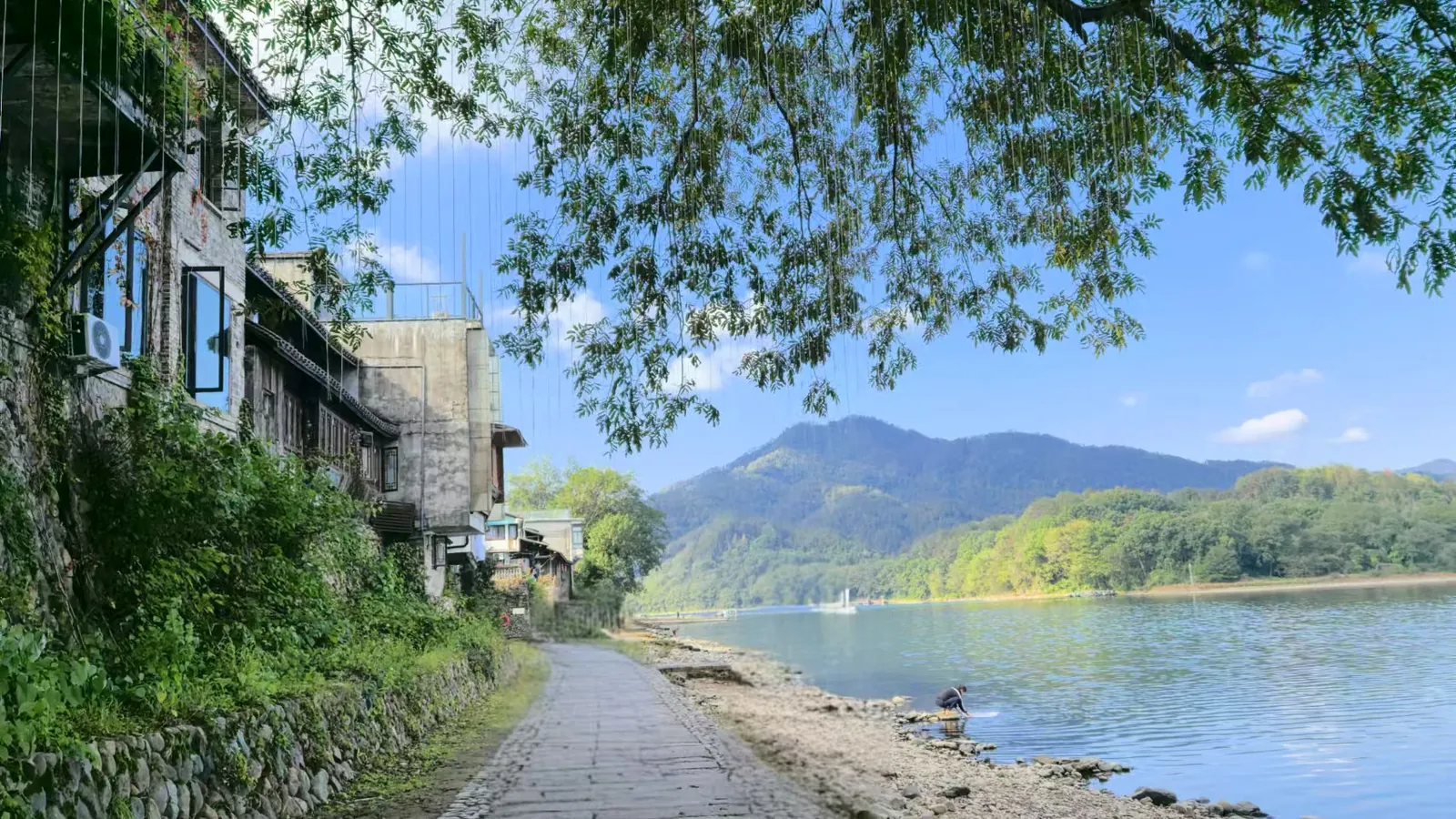 A paved waterside path with old two-storey buildings and climbing vines on the left, wooded hills across the water, and one person working at the water's edge