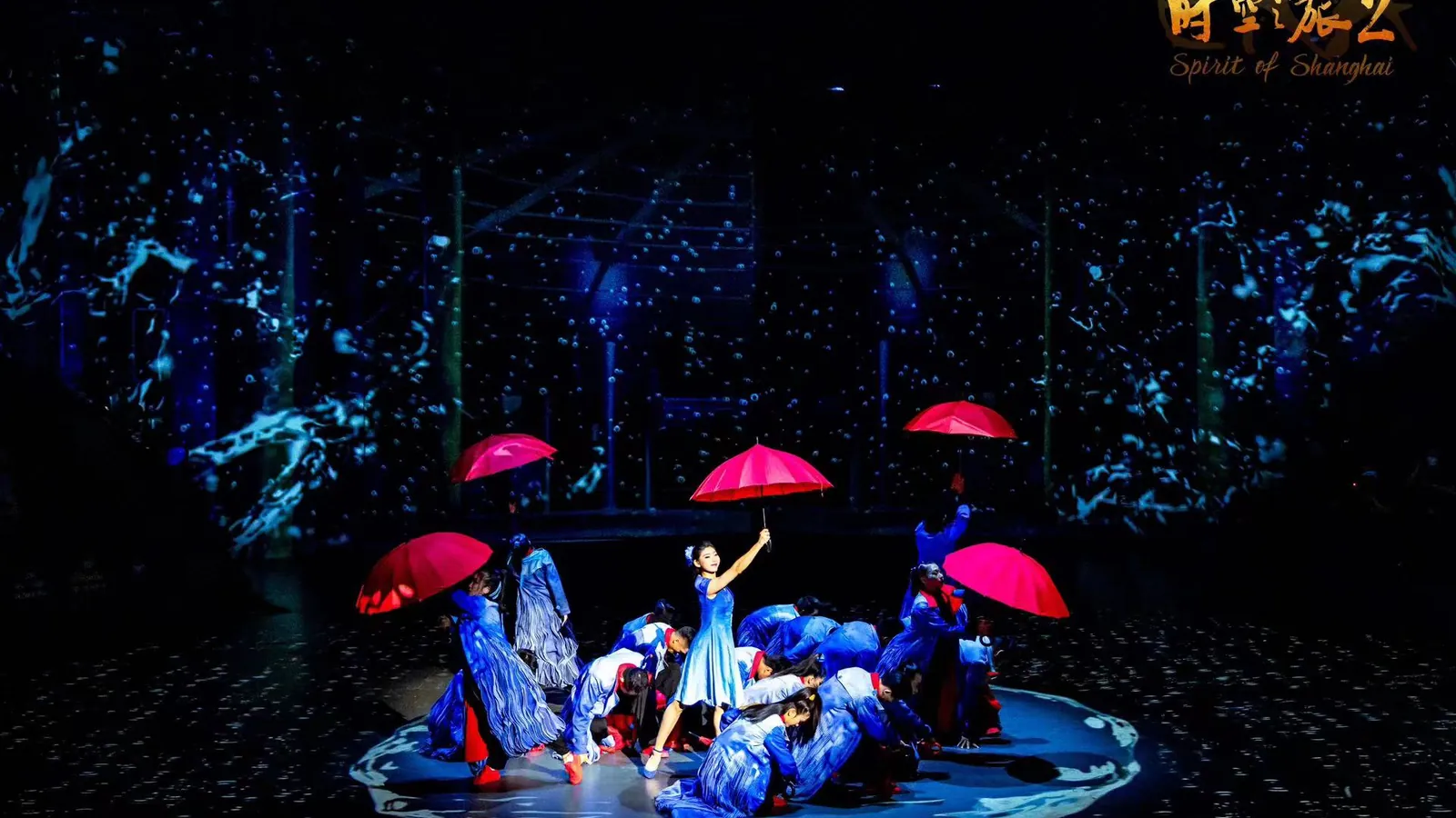 A dark stage in front of a blue projected backdrop, where a dancer in a pale blue dress raises a red umbrella above a group of performers in blue-and-white costumes crouching around her, with the show's logo in the corner of the frame