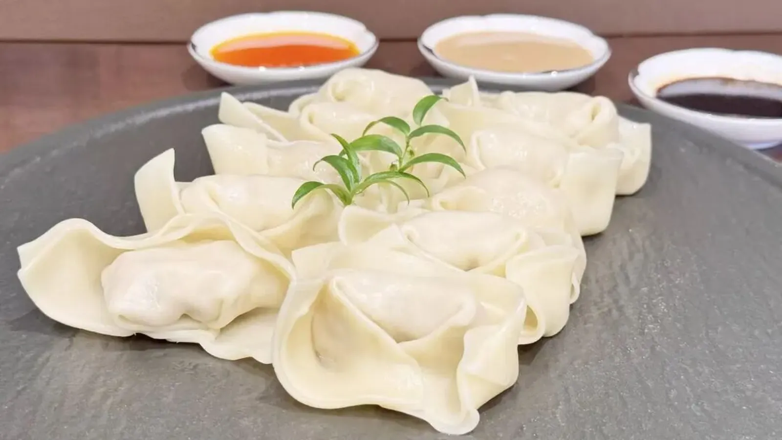 Around a dozen pale, thinly wrapped wontons on a grey stone platter, a single green sprig in the centre, with three small white dishes of chilli oil, pale sesame sauce and dark soy behind
