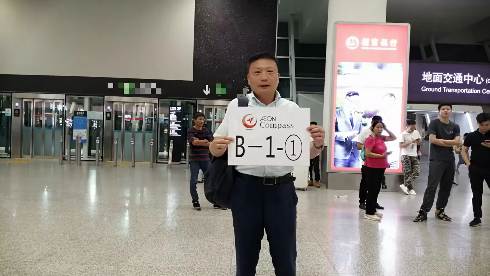 An airport arrivals hall with a man in a pale shirt holding up a large white placard printed with a meeting-point code, glass doors, passengers behind him and a Ground Transportation Centre sign overhead